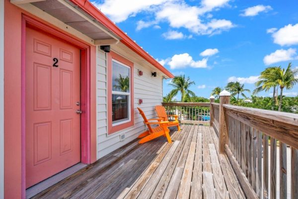 A wooden deck with orange chairs, a pink door numbered 2, railing, and a view of palm trees under a blue sky with white clouds.