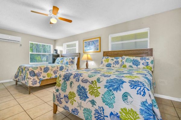 The image shows a bedroom with two beds covered in colorful floral bedding, a lamp, artwork, and windows, creating a bright and cozy atmosphere.