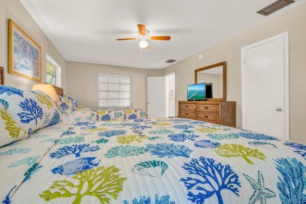 A bedroom with two beds featuring ocean-themed bedding, a ceiling fan, a dresser with a TV, and two artworks on the walls.