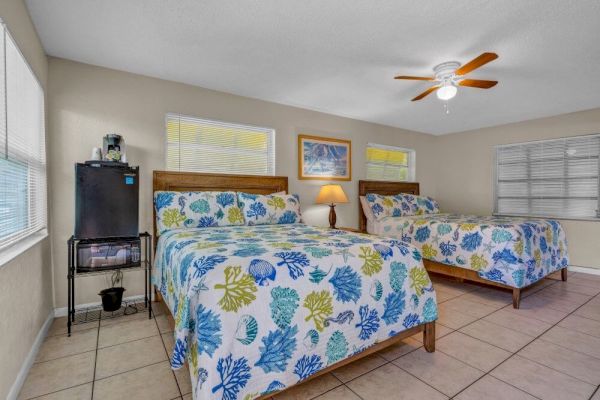 The image shows a room with two beds, floral bedspreads, a small fridge, a lamp, and a ceiling fan. Bright windows light the space.