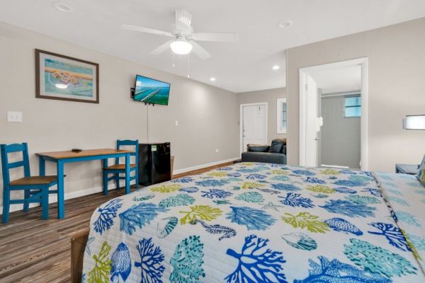 A bedroom with a colorful bedspread, a small table with chairs, a TV mounted on the wall, and a visible bathroom door.