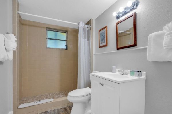 A bathroom with a shower, toilet, and sink. It includes a mirror, towels, toiletries, and a light fixture above the mirror.