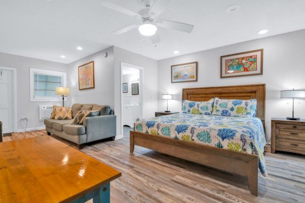 A bright bedroom with a colorful patterned bedspread, wooden furniture, a sofa, wall art, and ceiling fan. Modern and cozy atmosphere.