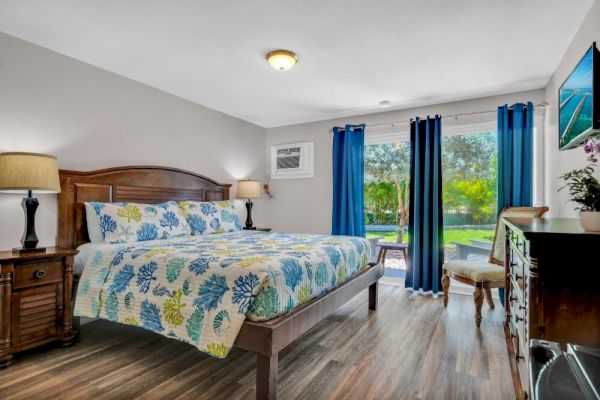 A bedroom with a floral-patterned bed, two nightstands with lamps, blue curtains, a dresser, and a window showing a garden view.