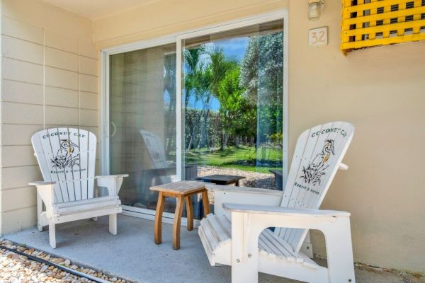 Two Adirondack chairs and a small table on a patio, near a sliding glass door. Lush greenery is reflected in the window, outside view.