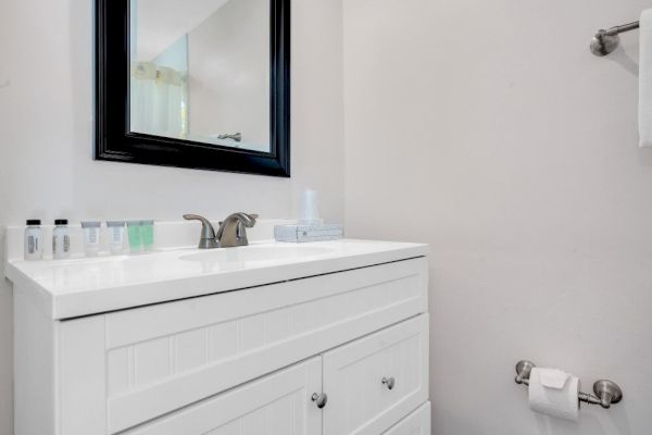 A white bathroom vanity with a mirror, toiletries, towel, and a toilet paper holder on the wall. The space is minimalist and tidy.