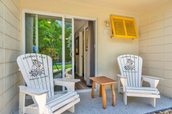 A cozy patio with two white Adirondack chairs and a small wooden table, set outside a sliding glass door with greenery beyond.