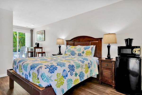 A cozy bedroom with a wooden bed, floral bedding, two lamps, a small table, chairs, and a mini fridge by the side.