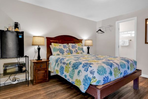 A cozy bedroom with a double bed, colorful bedding, two lamps, a mini fridge, and a small TV stand next to a closet.