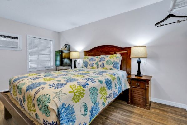 A cozy bedroom with a colorful sea-themed bedspread, two bedside lamps, a wooden nightstand, and a wall-mounted air conditioner.