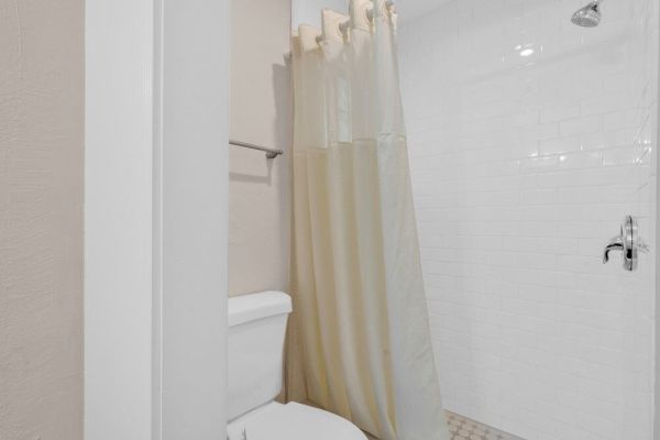 The image shows a bathroom with a toilet, a shower area with a white curtain, and white tiled walls.