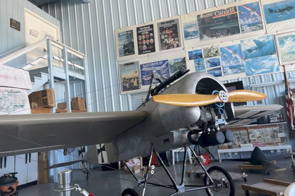 The image shows a vintage aircraft displayed in a museum, surrounded by aviation-themed posters on a corrugated metal wall.