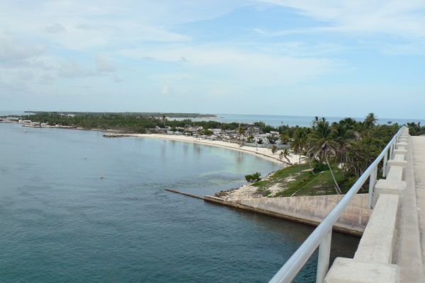 A coastal view from a bridge with clear blue water, lush green trees, and a small settlement by the shoreline under a partly cloudy sky.