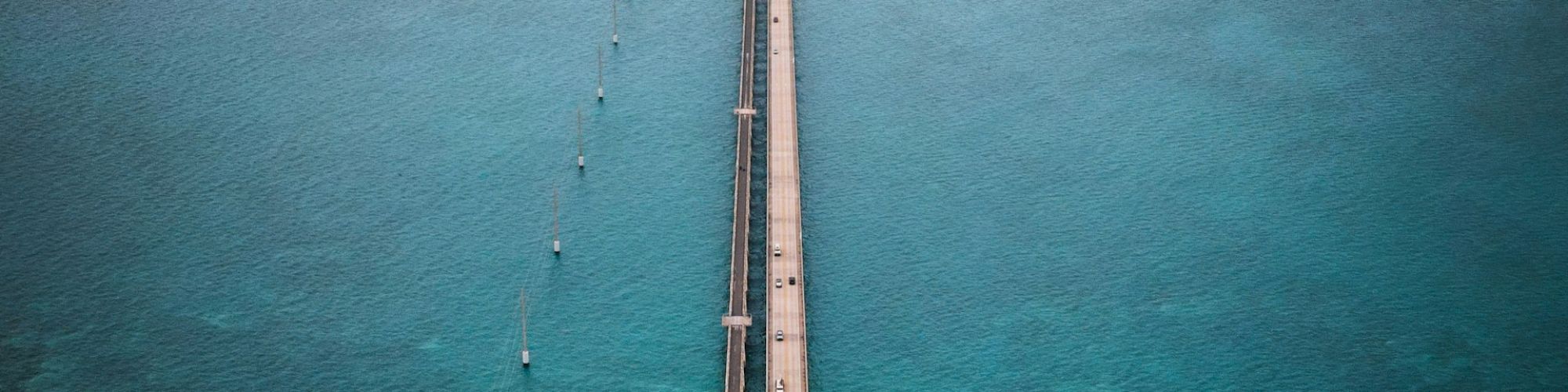 An aerial view of a long bridge stretching across turquoise waters, connecting landmasses, under a partly cloudy sky.