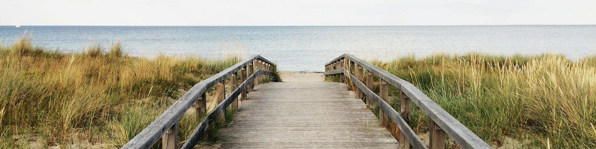 A wooden boardwalk leads through dunes to a calm sea under a partly cloudy sky.