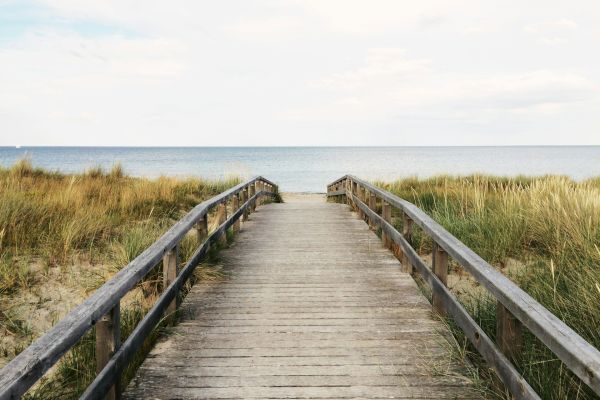 A wooden boardwalk leads through tall grass towards a beach with a calm sea and cloudy sky, creating a serene coastal view.