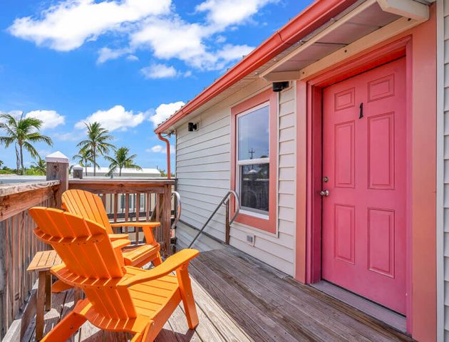 A sunlit deck with two orange chairs, a pink door, tropical palms, and a bright blue sky with clouds.