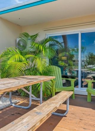 The image shows a patio with a wooden picnic table, colorful chairs, and sliding glass doors, surrounded by lush green plants and clear skies.