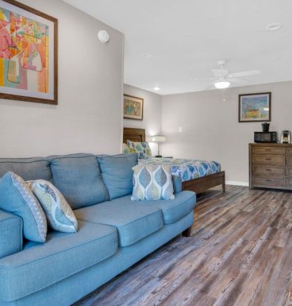 A cozy room with a blue sofa, artwork, a bed, and a dresser on wooden flooring. A TV is mounted on the wall, adding to the inviting space.