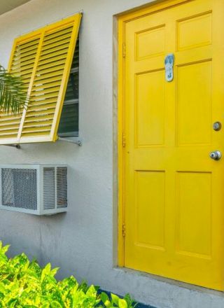 A white building features a bright yellow door and shutters, numbered 40, adjacent to vibrant green foliage and an AC unit.