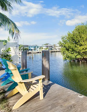 Two wooden adirondack chairs sit on a weathered dock by calm water, with palm trees and mangroves lining a sunny, tropical shoreline.
